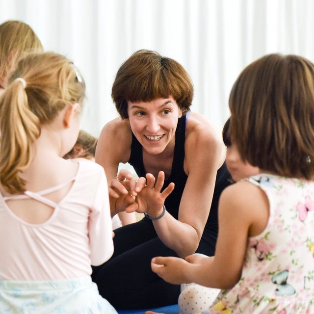Tanzlehrerin Afra Sentner hockt lachend im Kreis mit kleinen Kindern und zeigt ihnen eine Handbewegung